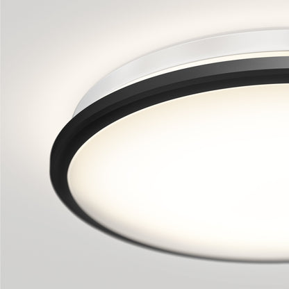 A close-up of the Artika-CAN-dev Vera 13 in. LED modern flush mount light features a matte black finish, silver rim, glowing white center emitting 200 lumens, and is mounted on a white ceiling.