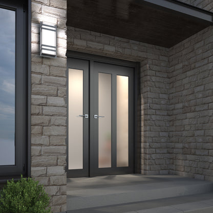 Modern double entry doors with frosted glass panels set in a stone exterior wall. A rectangular wall light is mounted beside the doors, and a small plant is visible in the lower left corner.