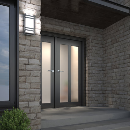 A modern house entrance features double frosted glass doors in a stone wall. To the left of the doors, the Artika Glowbox 3 indoor-outdoor LED wall light is mounted, with a small green bush visible in the lower left corner.