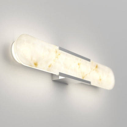 The Artika Selena 3 CCT LED bathroom vanity light features a frosted, marble-like glass cover and a silver rectangular base, emitting a soft, warm glow when mounted on a light gray wall.