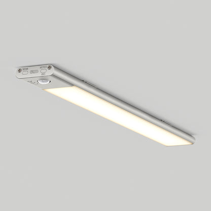 The Artika Outline under cabinet LED light 3CCT is a slim, rectangular fixture with a silver metallic frame and soft white illumination, featuring visible control buttons and mounted on a plain light gray ceiling.
