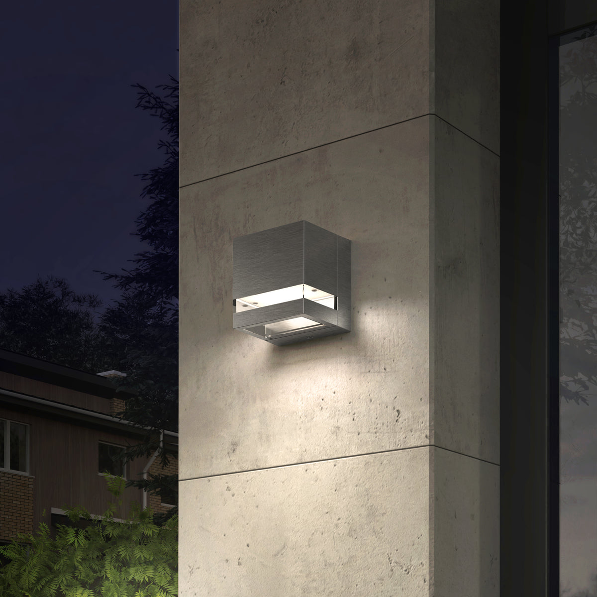 The Artika Valor Square LED indoor-outdoor wall light is mounted on a concrete pillar, casting soft white light at night. Trees and a building in the background enhance the modern outdoor lighting ambiance.