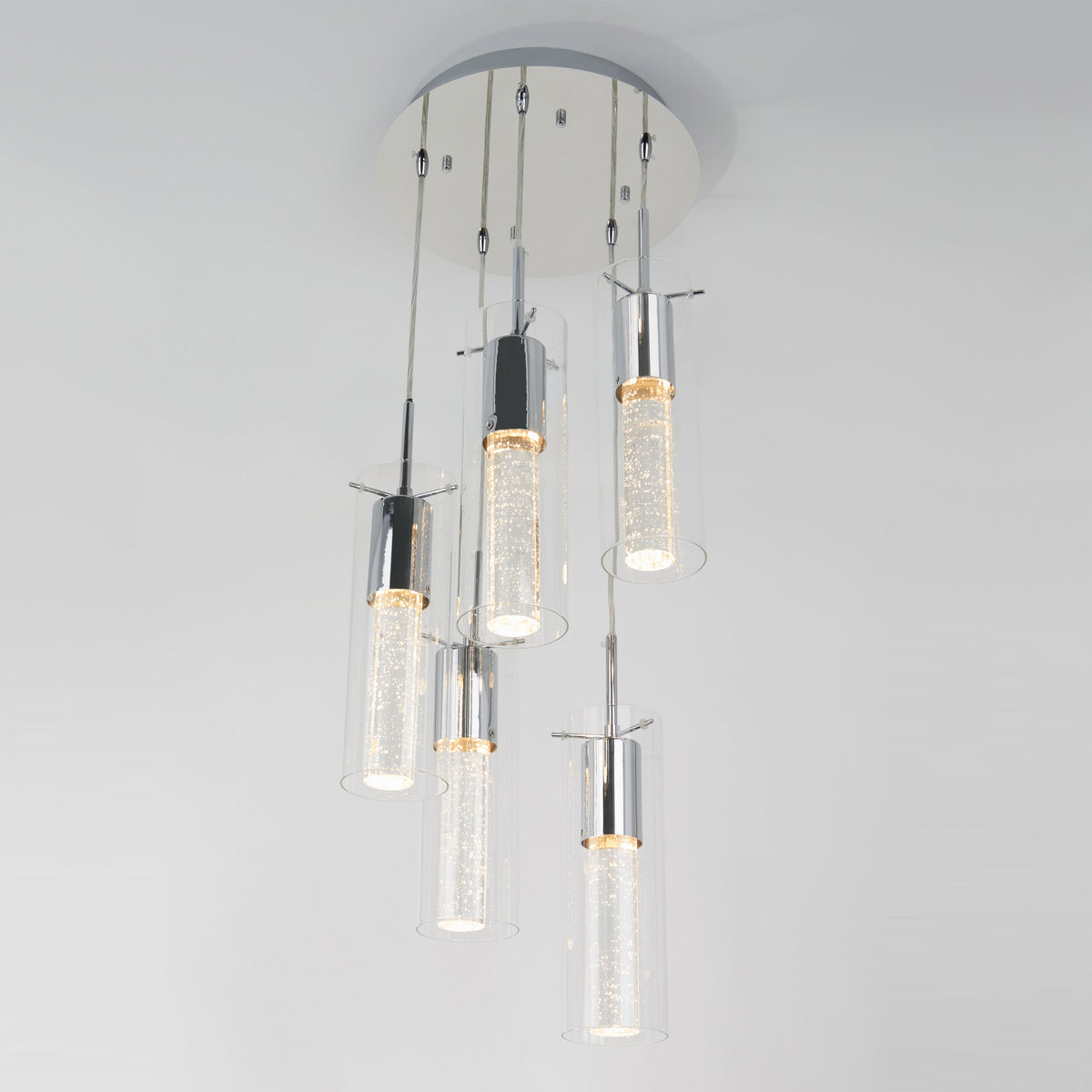 A modern ceiling light fixture with five cylindrical glass pendants, each containing bubbles and suspended at different lengths from a round white base, emits a soft white glow.