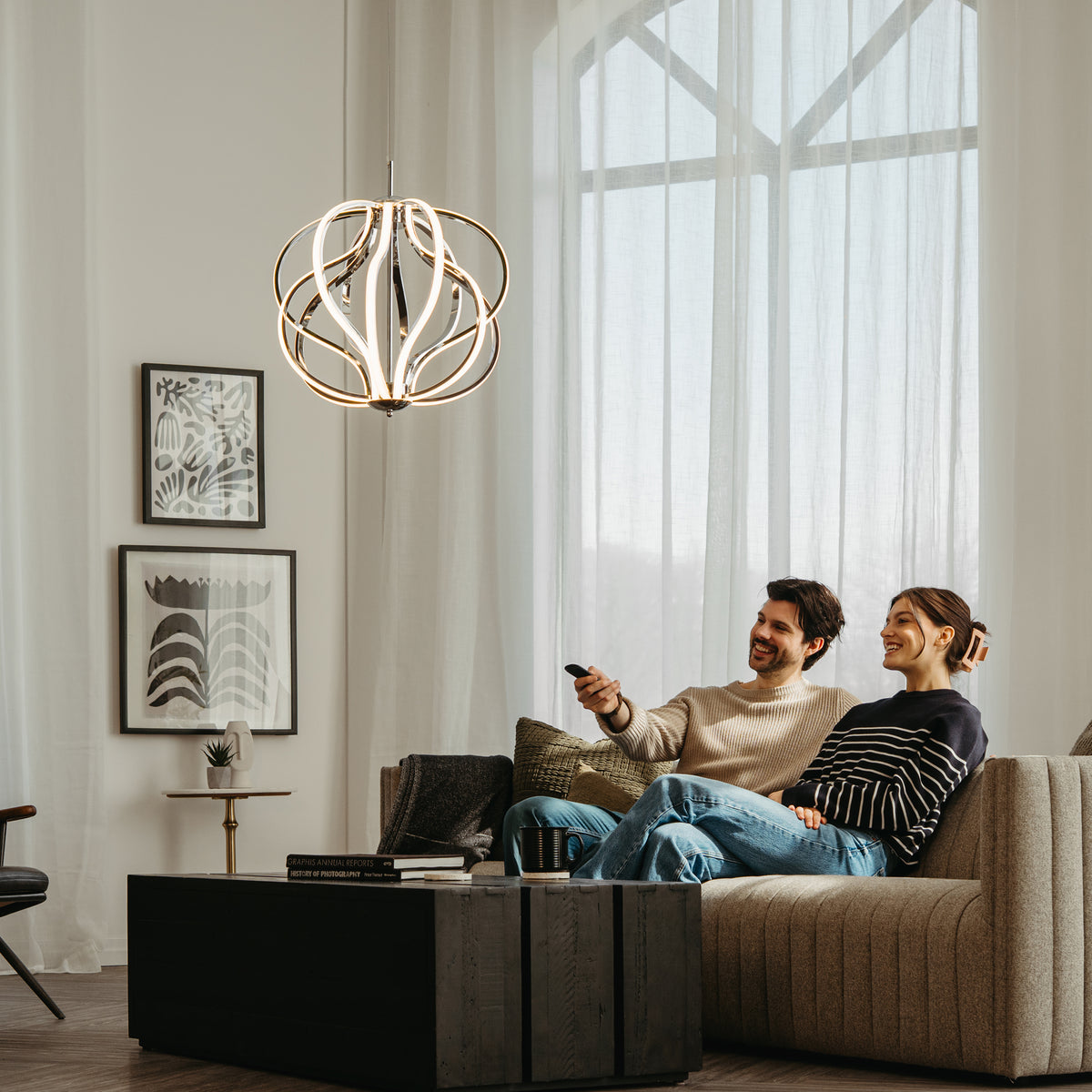 A smiling couple sits on a beige couch in a modern living room, watching TV. They are relaxed, with the man holding a remote. Large windows, art on the wall, and a contemporary chandelier decorate the space.