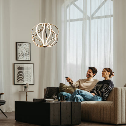 A smiling couple sits on a modern beige sofa in a bright living room, watching TV. Large windows, abstract wall art, a black coffee table, and the Artika Meridian LED pendant light add style to the space.