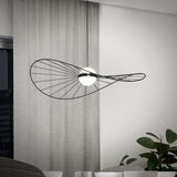 A modern pendant light with a sculptural, black wire frame and two white globes hangs in a minimalist room with light gray curtains, a window, and a green potted plant.