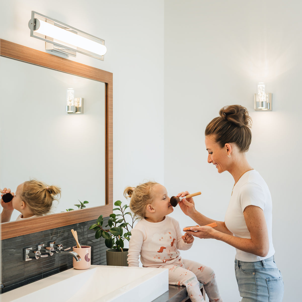 A woman smiles, applying makeup to a girl by an Artika Rialto 27-in LED Vanity Light Bar, near a mirror and potted plant.
