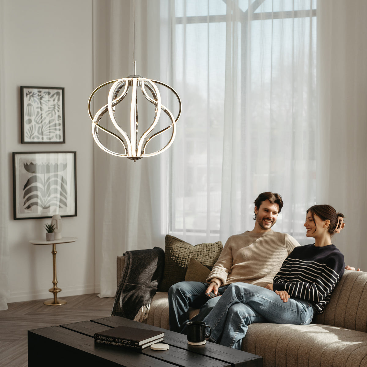 A couple sits smiling together on a modern sofa in a bright, stylish living room with large sheer curtains, framed art on the wall, and a contemporary hanging light fixture overhead.