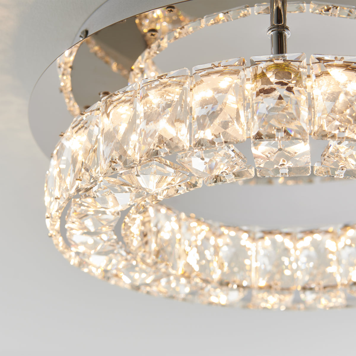 Close-up of a modern ceiling light fixture featuring two circular rings adorned with faceted crystals, emitting a warm, sparkling glow against a neutral background.