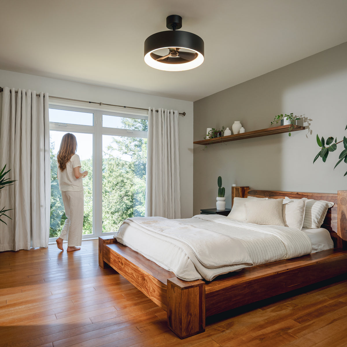 A woman stands in a bright room with plants, wooden floors, and an Artika Aiden 24 in. LED modern fandelier ceiling fan overhead.
