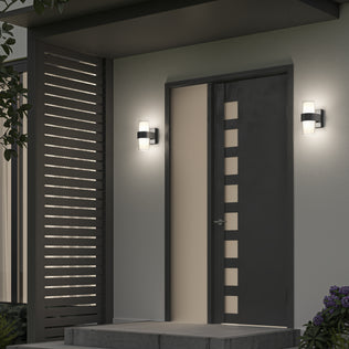 A modern front door with vertical rectangular cutouts is flanked by two Artika Colton modern LED indoor-outdoor wall lights (5 CCT). To the left is a slatted privacy screen, steps lead to the entrance, and plants are visible nearby.