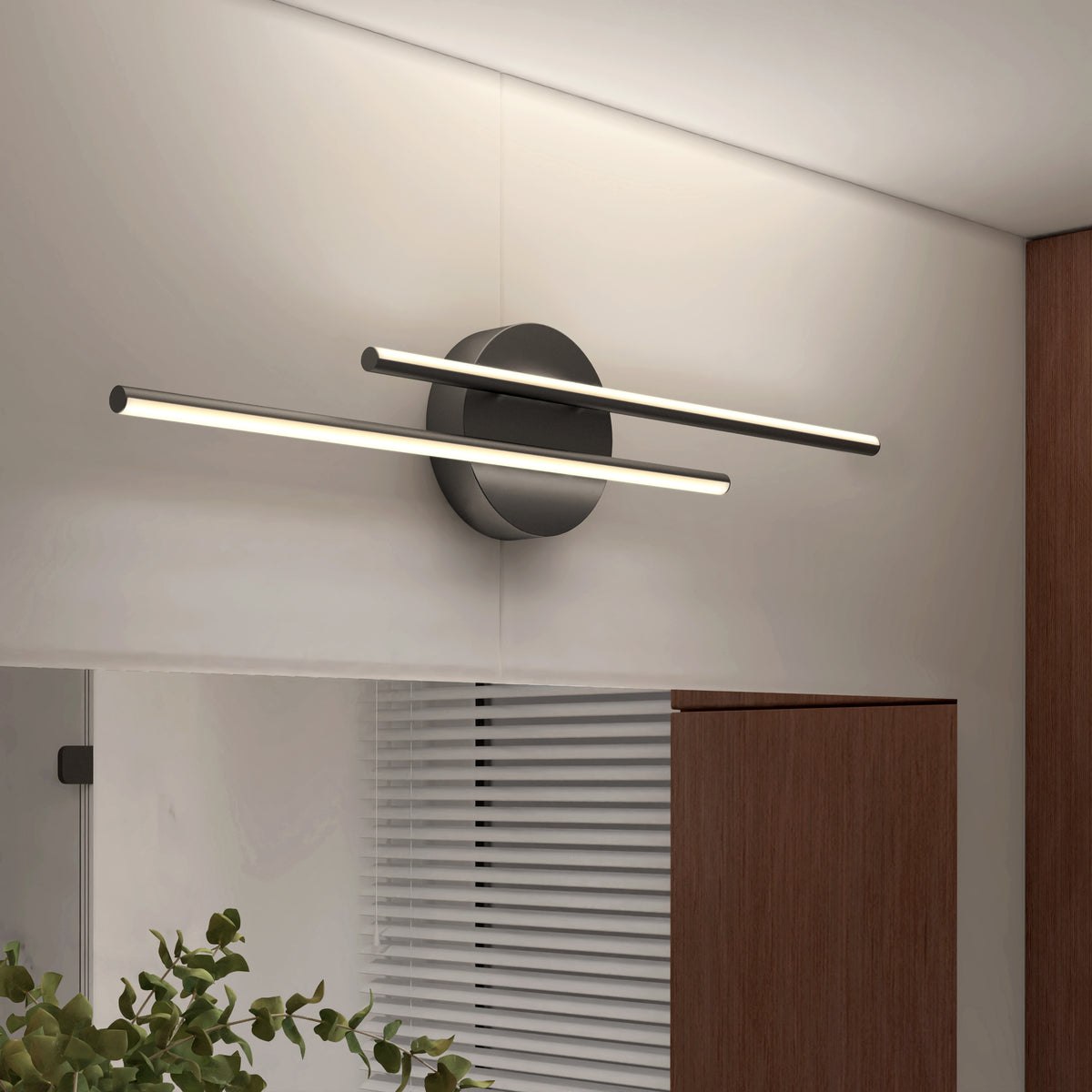 A modern wall light fixture with two parallel LED bars mounted on a round black base, installed above a mirror in a contemporary room with wooden cabinetry and white walls.