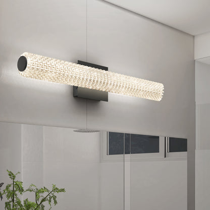 The Artika Exalto LED bathroom vanity light, featuring a modern cylindrical design with a textured, glowing glass surface, is mounted above a large mirror in a bright, minimalist bathroom with a small green plant in the corner.