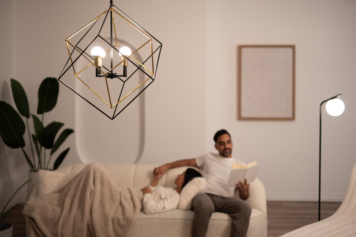 A modern geometric chandelier hangs in a cozy, softly lit living room. A couple relaxes on a white sofa; one person reads a book while the other rests with a blanket. A plant and a floor lamp decorate the space.